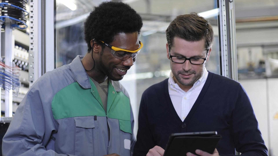 A male employee wearing safety clothing discussing with a man holding a digital tablet inside a factory